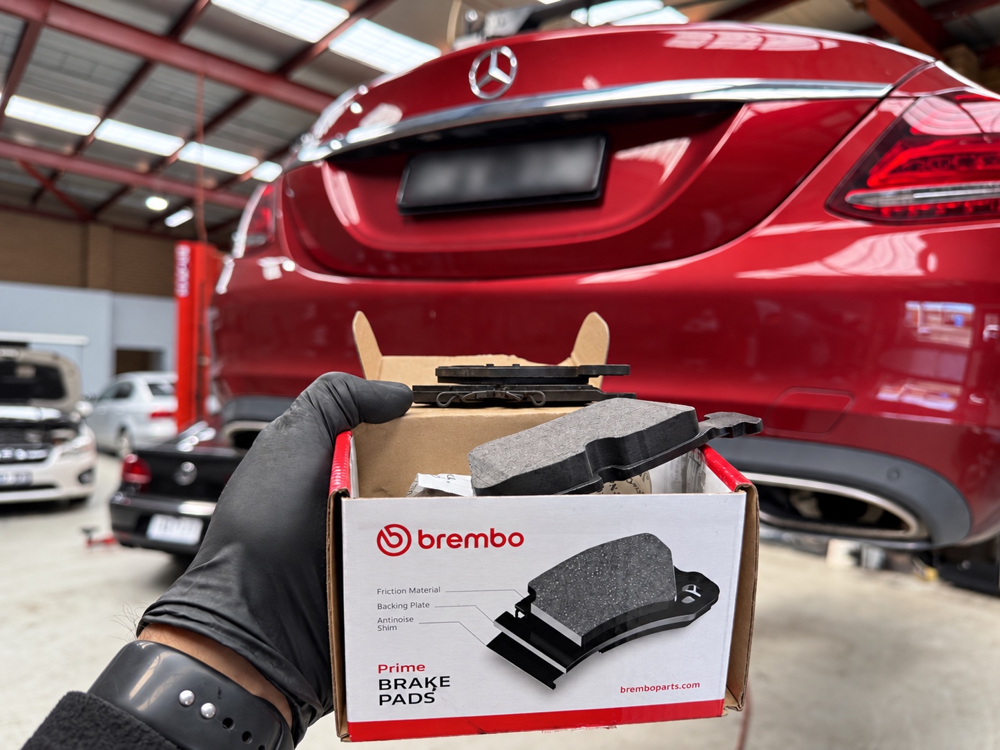 Brembo Prime rear brake pads held in front of a red Mercedes-Benz at the Luca Motors workshop.